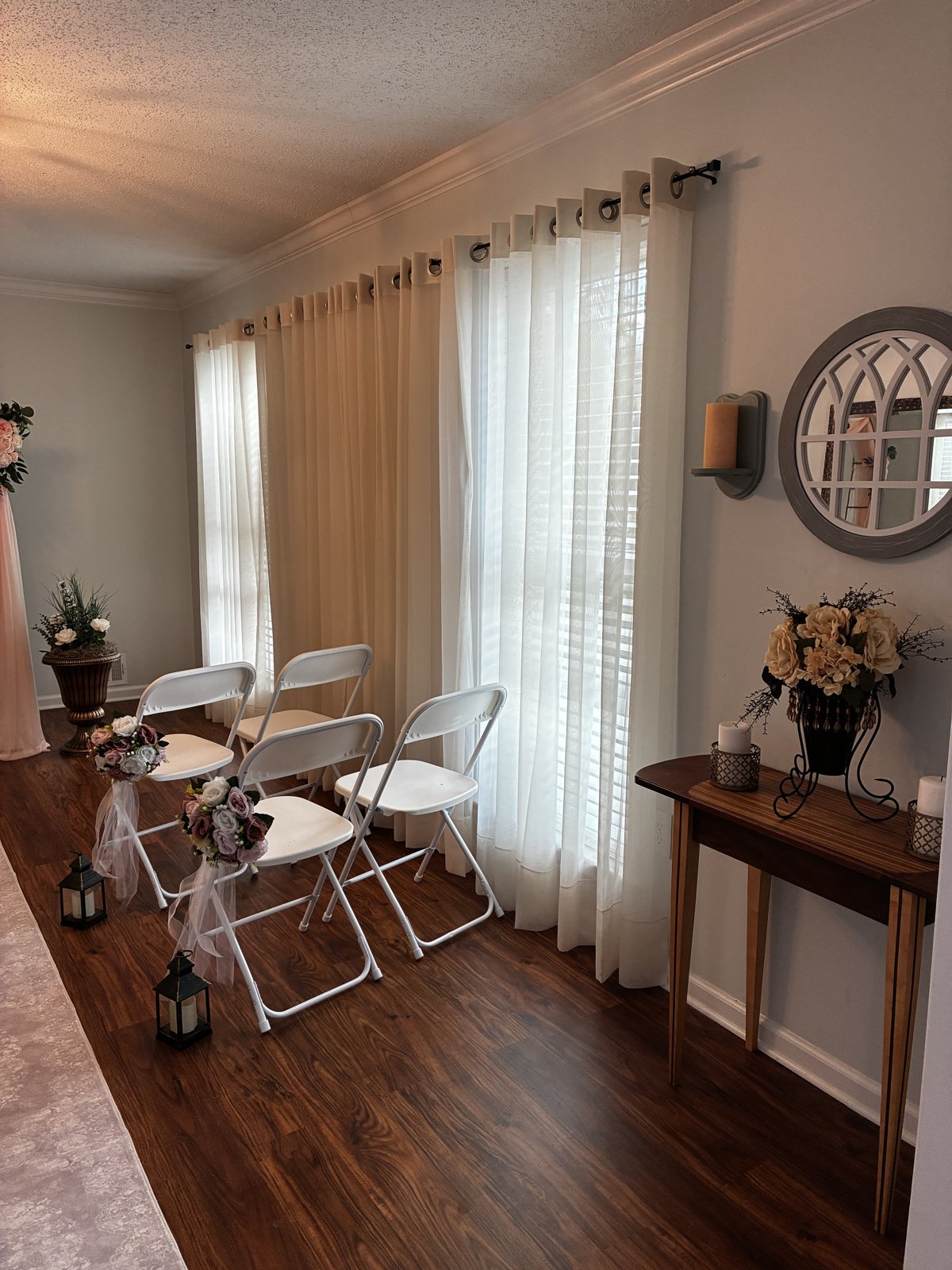 Guest seating beside tall windows with sheer curtains, flower bouquets and lanterns on the aisle chairs, and a candlelit side table beneath a round mirror